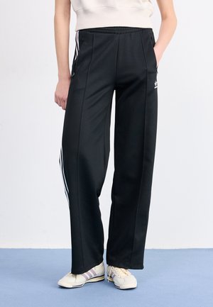 Person, die schwarze Adidas-Trainingshose mit weißen Streifen und weißen Sneakers trägt, steht mit den Händen in den Taschen auf blauem Boden.