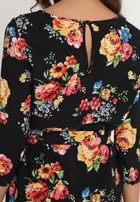 La robe fleurie présente un fond noir avec des roses multicolores et des fleurs plus petites, un dos en forme de clé et une taille cintrée.
