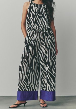 Frau trägt einen ärmellosen Zebra-Print-Jumpsuit mit weitem Bein und lila Bündchen, kombiniert mit schwarzen offenen Sandalen mit Absatz.