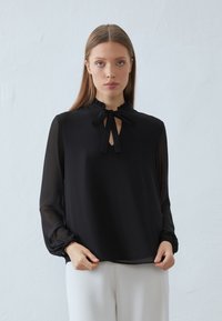 Zwarte blouse met een gerafelde kraag en een strik bij de hals. Lange, transparante mouwen en een losse pasvorm. Eenvoudig, elegant ontwerp zonder patronen.