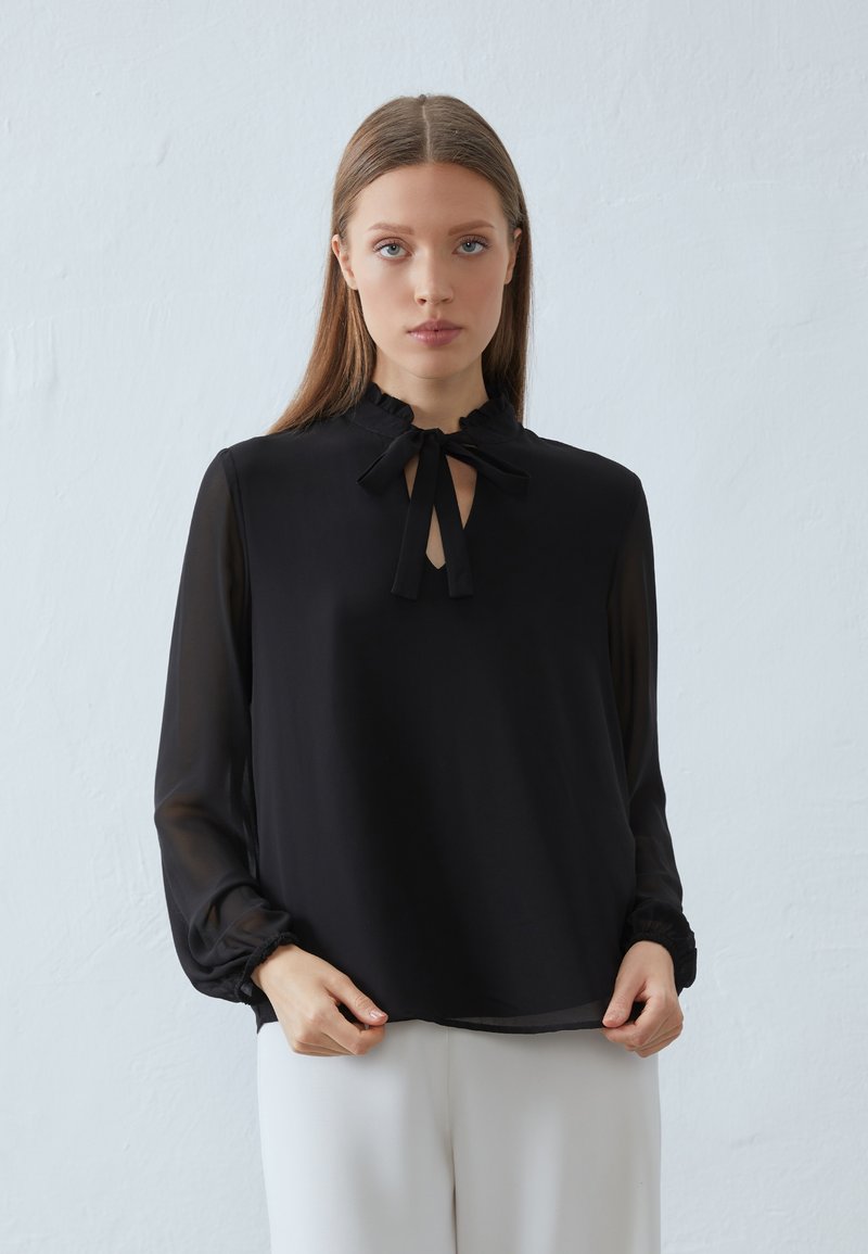 Zwarte blouse met een gerafelde kraag en een strik bij de hals. Lange, transparante mouwen en een losse pasvorm. Eenvoudig, elegant ontwerp zonder patronen.