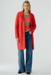 Manteau en mélange de laine rouge avec une fermeture à un bouton, associé à un pull rayé gris et corail et un jean en denim évasé.