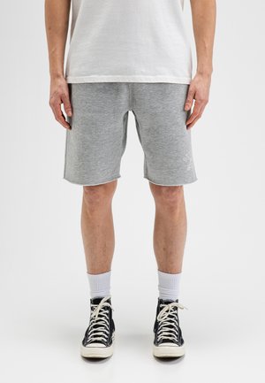 Mann trägt hellgraue lässige Shorts, weiße Socken, schwarze High-Top-Sneaker und ein schlichtes weißes T-Shirt und steht vor weißem Hintergrund.