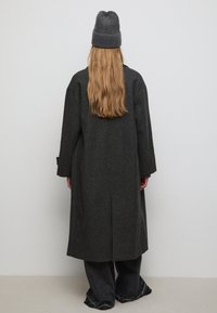 Lungo cappotto in lana grigio scuro con vestibilità rilassata, spalle scese e taglio dritto, abbinato a jeans di denim neri a gamba larga e un berretto di maglia grigia.