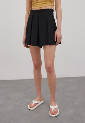 Woman wearing black pleated skirt shorts, mustard ruched crop top, and white flip-flop sandals standing on gray floor.
