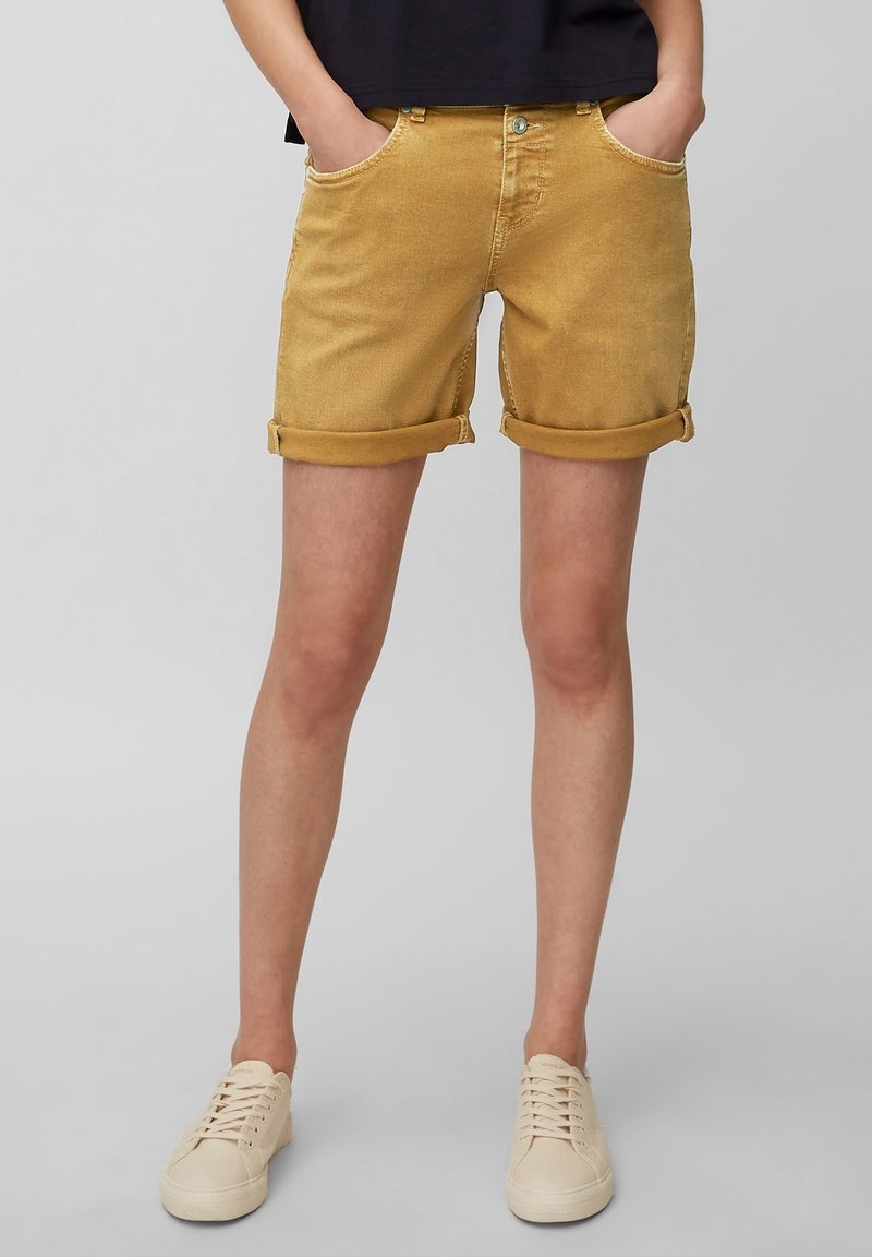 Personne portant un short moutarde retroussé et des baskets à lacets blanc cassé, debout les mains dans les poches devant un fond gris.