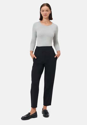 Maglia grigia a maniche lunghe abbinata a pantaloni neri con vita elasticizzata, tasche laterali e lunghezza corta, completata da mocassini neri. Design minimalista.