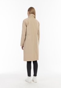 Beige dlouhý kabát s vysokým límcem, staženým pasem a dlouhými rukávy. Hladká textura a minimalistický design, zobrazeno zezadu s černými legínami.