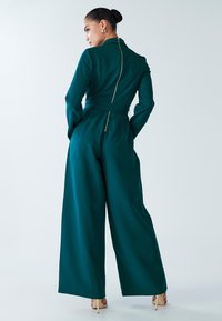Teal jumpsuit featuring a high collar, long sleeves, belted waist, and a visible gold zipper at the back. Wide-leg design with smooth texture.