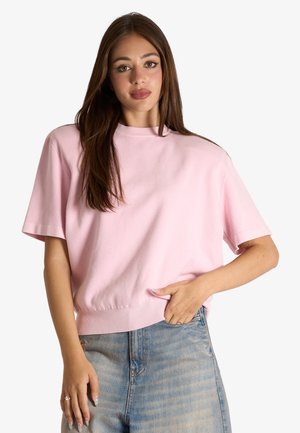 Donna con lunghi capelli castani che indossa una maglietta rosa chiaro a maniche corte e jeans blu, in piedi con una mano nella tasca.