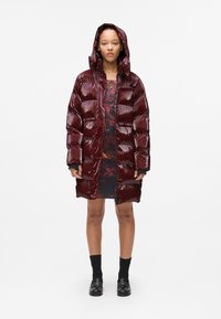 Shiny, maroon puffer coat with a hood, featuring a quilted design and front zipper. Worn over a black and red patterned dress.