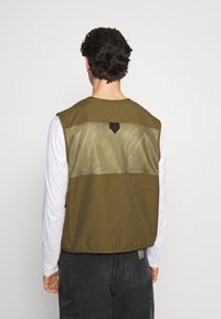 Man som bär en olivgrön ärmlös väst med nätpanel över en vit långärmad skjorta och mörka cargo-byxor, vänd bort från kameran.