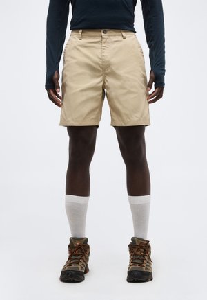 Άντρας που φοράει μπεζ σορτς, λευκές κάλτσες, ορειβατικά παπούτσια και μακρυμάνικο σκοτεινό πουκάμισο, στέκεται μπροστά σε ένα απλό φόντο.