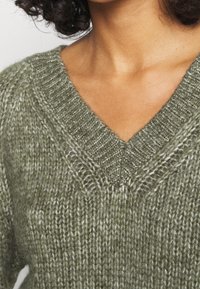 Olivgrön stickad tröja med V-ringning, med texturerad garn och ribbad hals. Närbild på tygdetalj.