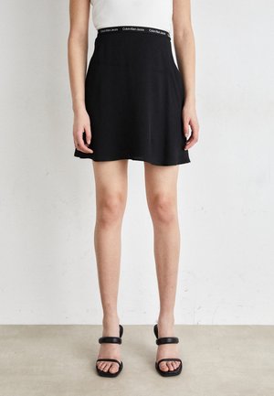 Woman wearing a black mini skirt with Calvin Klein Jeans elastic waistband and black open-toe heeled sandals, standing against a plain wall.