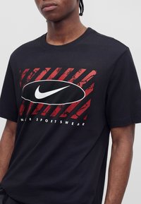 Νέος άνδρας που φοράει μαύρο μπλουζάκι Nike με κόκκινες διαγώνιες ρίγες και λευκό λογότυπο Nike swoosh μέσα σε μαύρο οβάλ.