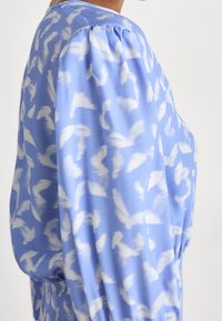 Světle modrá blůza s bílým abstraktním vzorem, se zdůrazněnými rukávy a mírně volným střihem. Měkký materiál s hladkou texturou.