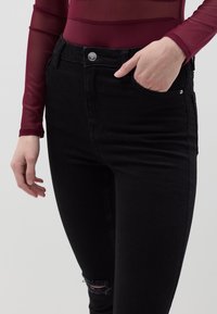 Vaqueros de mezclilla negra de corte ajustado, con bolsillos frontales, cierre de botón plateado y un detalle de rodilla desgastada. El material parece resistente.