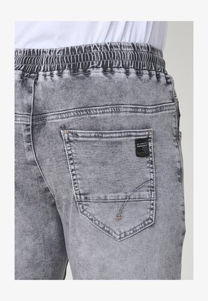 Szürke farmernadrágos joggerek, elasztikus derékgumival és hátul egy varrott dizájnnal ellátott zsebbel, valamint egy címkével. A textúra sima, elhalványult felülettel.