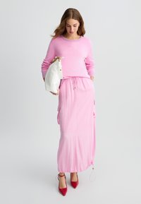 Maglione rosa con collo a costine e dettagli ricamati, abbinato a una lunga gonna rosa con vita regolabile e tasche laterali. Tacchi rossi e borsa bianca.