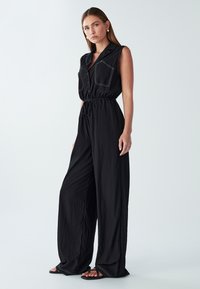 Zwarte jumpsuit met korte mouwen, knoopsluiting aan de voorkant, twee borstzakken en een elastische trekkoordtaille. Heeft contrasterende witte stiksels.