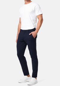 Pantaloni slim-fit blu navy con una texture liscia, dotati di tasche laterali, passanti per cintura e chiusura con bottone, abbinati a una t-shirt bianca semplice e sneakers bianche.
