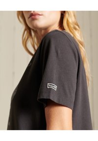 Camiseta negra de algodón con mangas cortas, que cuenta con un parche de logotipo en la manga izquierda. Textura suave y corte recto.