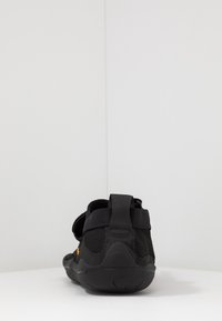 Chaussures de sport noires avec partie supérieure en mesh, sangle ajustable et semelle en caoutchouc texturée. La vue arrière montre le tirant au talon et le détail de marque.