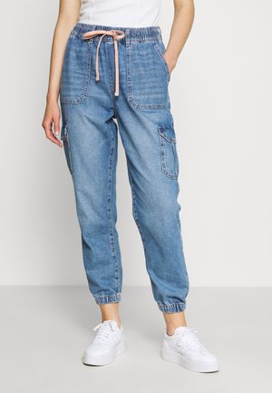Personne portant un pantalon jogger en denim bleu avec de grandes poches avant et latérales, des chevilles élastiques, une taille à cordon rose et des baskets blanches.