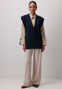 Námornícky strihový vestový kabát s ramennými vycpávkami cez béžovú blúzku, s predným zapínaním na gombíky, širokými rukávmi a s krémovými nohavicami s voľným strihom.