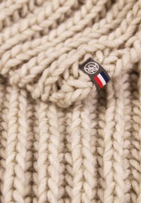 Gebreide crèmekleurige sjaal van dikke, gestructureerde garen; voorzien van een klein label met een logo en de kleuren van de Franse vlag.