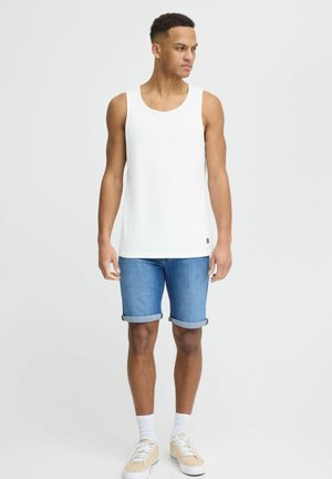 Weißes Baumwoll-Top kombiniert mit gerollten Denim-Shorts. Das Modell trägt weiße Socken und hellbraune Sneakers. Schlichtes Design, lässige Passform.