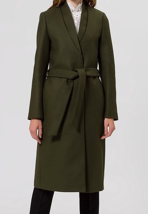 Kobieta w długim, oliwkowozielonym płaszczu z paskiem, nałożonym na biały koronkowy top i czarne spodnie, stojąca na tle jasnoszarego tła.