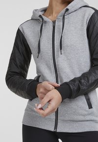 Siva zip-up dukserica s preklopnim crnim umjetnim kožnim rukavima, kapuljačom na vezanje i rebrastim manžetama. Glatka tekstura s kontrastnim materijalima.
