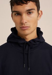 Jūrinės mėlynos spalvos džemperis su raišteliu ant kaklo, minkšta tekstūra ir matomais metaliniais akcentais. Paprastas, traukiamas dizainas.