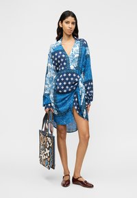 Μπλε φόρεμα με σχέδιο, μακριά μανίκια και σχεδιασμό τύπου τύλιγμα, με διάφορες αποχρώσεις του μπλε και περίπλοκες λεπτομέρειες. Συνοδεύεται από τσάντα totes με σχέδιο.