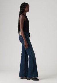 Nainen, jolla on pitkät letitetyt hiukset, pukeutuneena mustaan hihattomaan puseroon ja tummansiniset leveälahkeisiin farkkuihin, seisoo sivuprofiilissa yksinkertaista harmaata taustaa vasten.