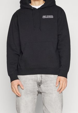 Czarna bluza z kapturem i kieszenią z przodu, z białym logo "OBEY STUDIOS". Materiał bawełniany, luźny krój, regulowany kaptur ze sznurkiem.