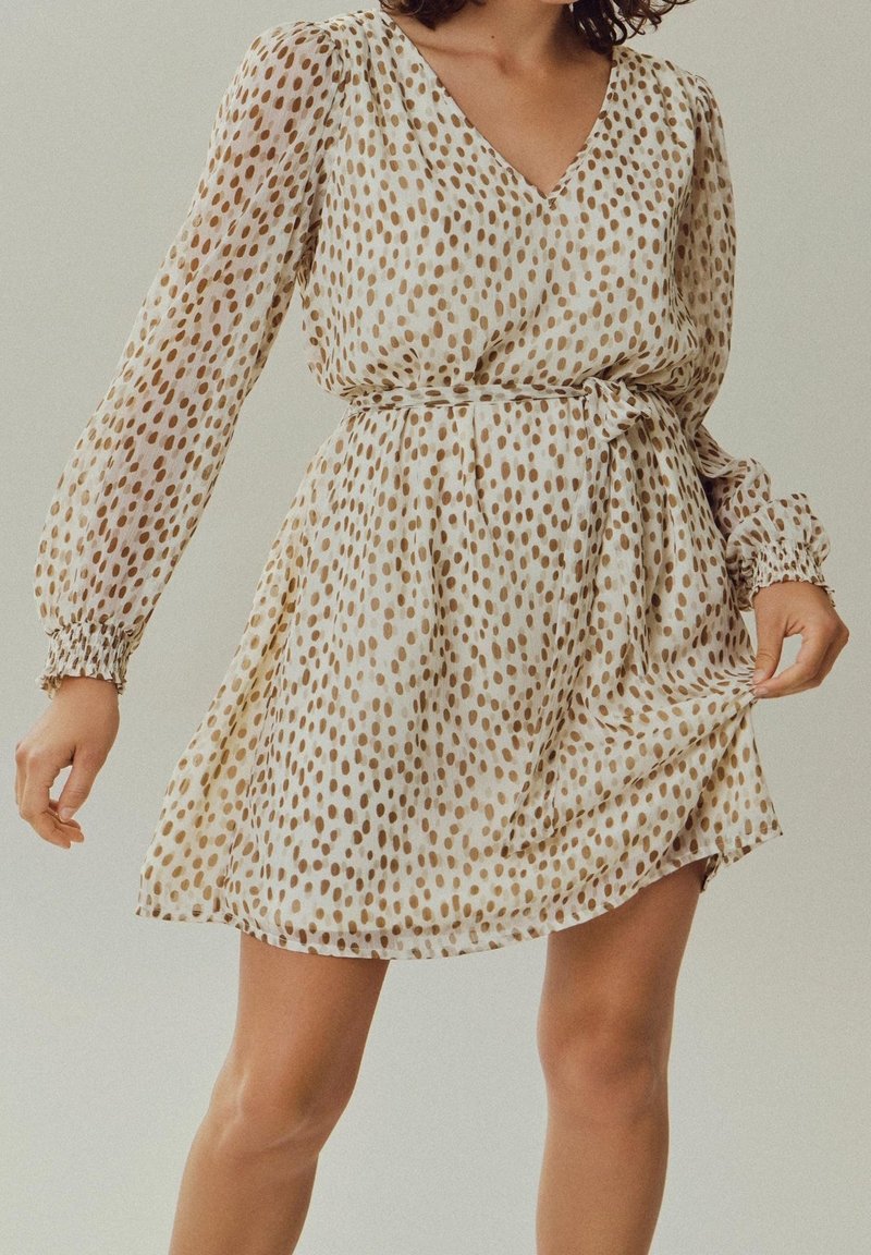 Femme portant une robe beige à manches longues avec des pois marron, tenant l'ourlet de la robe d'une main, taille ceinturée d'une ceinture en tissu.