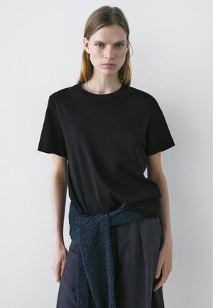 Kvinde med glat blondt hår iført en ensfarvet sort T-shirt med korte ærmer og mørke bukser med en blå strikket sweater bundet omkring taljen.