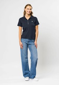 Navy polo shirt med korte ermer, tre-knaps placket, og en liten logo. Kombinert med lyseblå vide jeans og hvite joggesko.