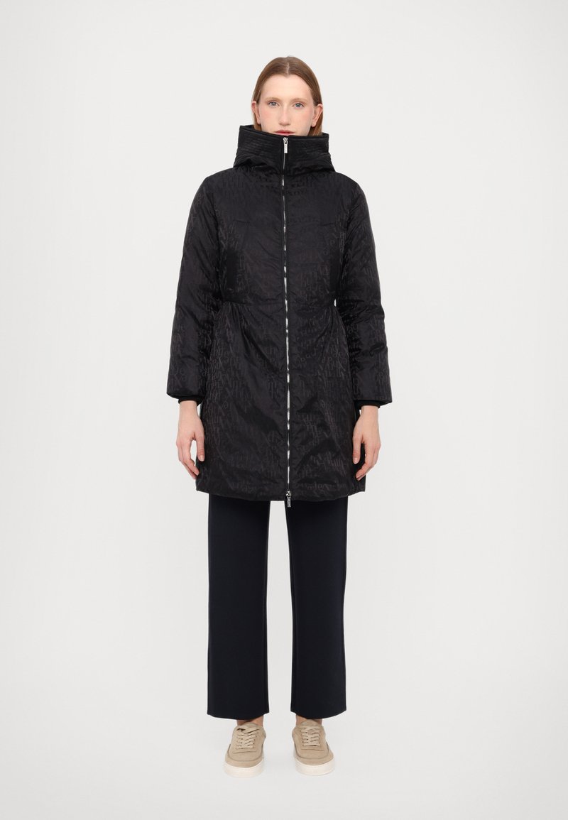 Giacca a vento nera trapuntata con colletto alto, zip frontale e due tasche laterali. Abbinata a pantaloni scuri e sneakers beige.