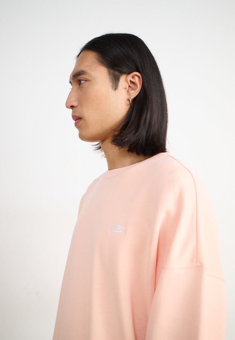 Sweatshirt surdimensionné rose clair avec un logo Nike blanc sur la poitrine, featuring une texture lisse et un col rond, présenté sur un modèle.