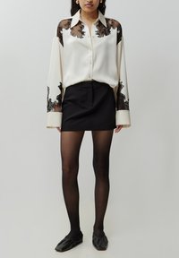 Witte satijnen blouse met zwarte kantdetails op de schouders en cuffs, gecombineerd met een zwarte minirok en sheer zwarte panty’s. Zwarte platte schoenen.