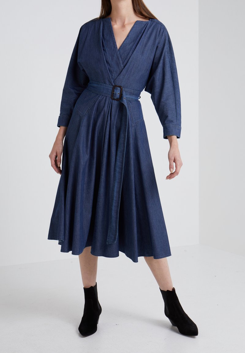 Femme portant une robe midi en denim bleu à manches longues avec une ceinture assortie et des bottines noires, debout devant un fond blanc uni.