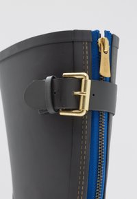 Bota de goma negra con una hebilla dorada, cremallera azul y detalles de costura amarilla. El diseño presenta una textura suave y una forma alta.