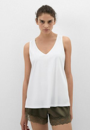 Kobieta ubrana w białą, bezrękawną bluzkę z dekoltem w serek oraz oliwkowozielone szorty z ozdobną koronką, stojąca na jednolitym jasnoszarym tle.