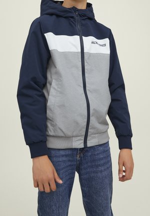 Person trägt eine blau-weiße-graue Kapuzenjacke mit Reißverschluss und blaue Jeans, steht mit entspannten Armen seitlich.