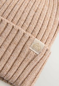 Beanie en tricot côtelé beige avec une finition texturée et une étiquette crème cousue présentant un design de logo subtil.