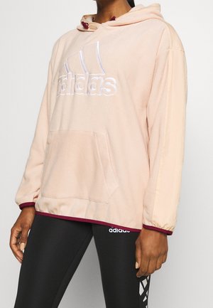 Persoon draagt lichtroze Adidas hoodie met voorzak en zwarte Adidas legging met wit logo op de dij, staand tegen een effen achtergrond.